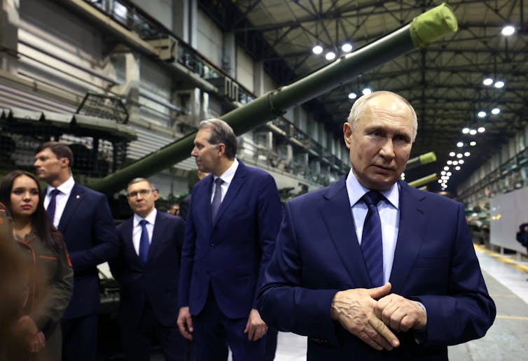 A man in a suit stand sin front of a tank.