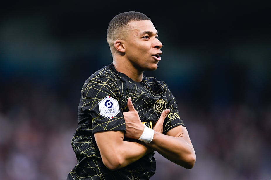 Kylian Mbappé wearing a black football jersey with his arms crossed.