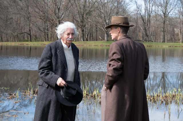 Un hombre de bigote y pelo blanco habla con otro, ambos con abrigos largos, delante de un lago.