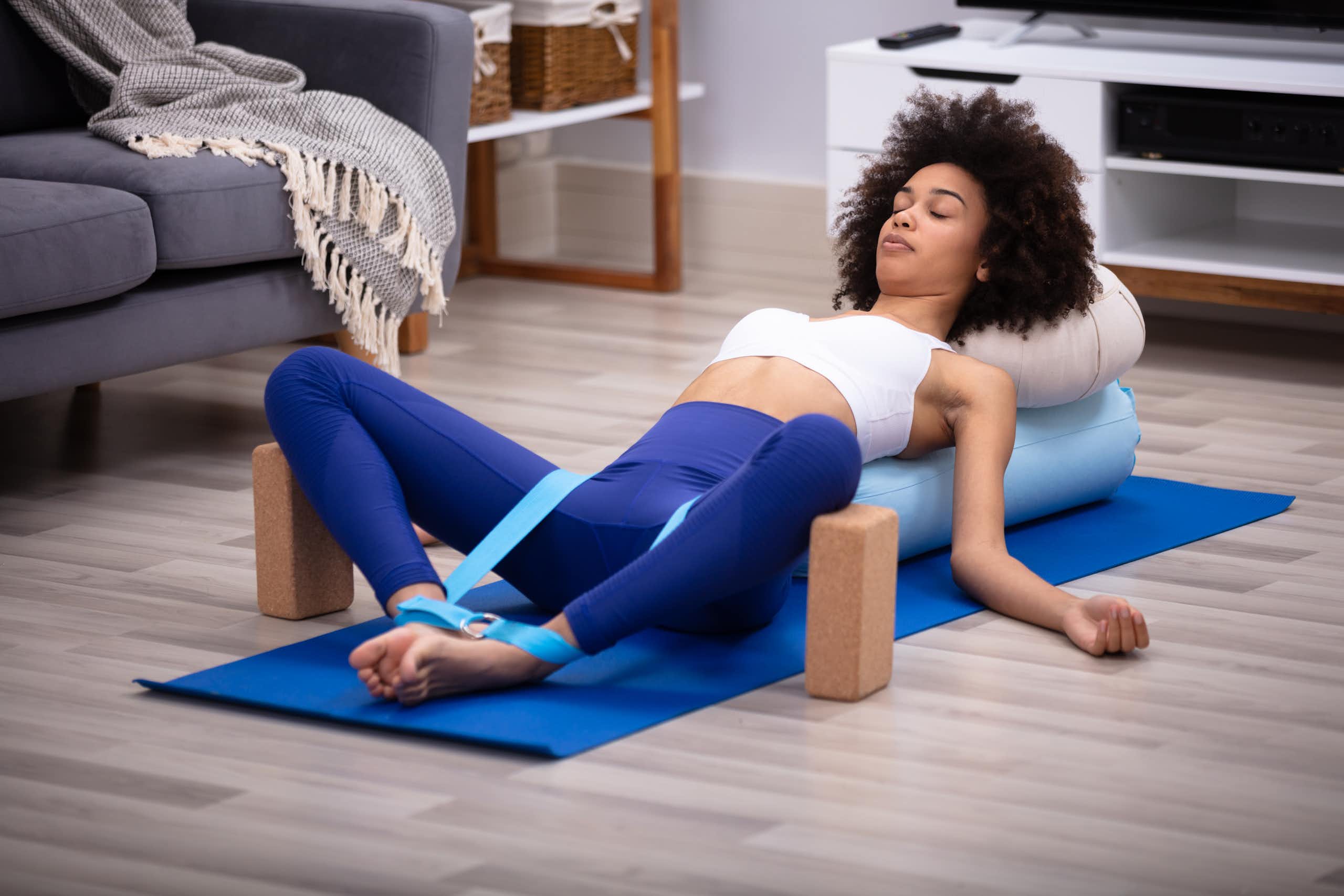 Une jeune femme est allongée sur le dos sur un tapis de gymnastique, les yeux fermés, les jambes en position de yoga (jambes écartées mais pieds joints, le haut du corps reposant sur des coussins.