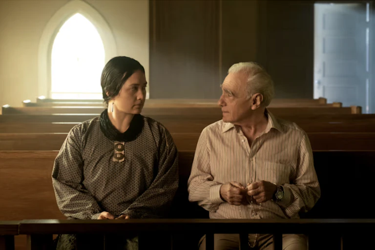 A dark-haired woman in a dark dress and a white-haired man in a button-down shirt sit looking at each other seriously in a church pew.