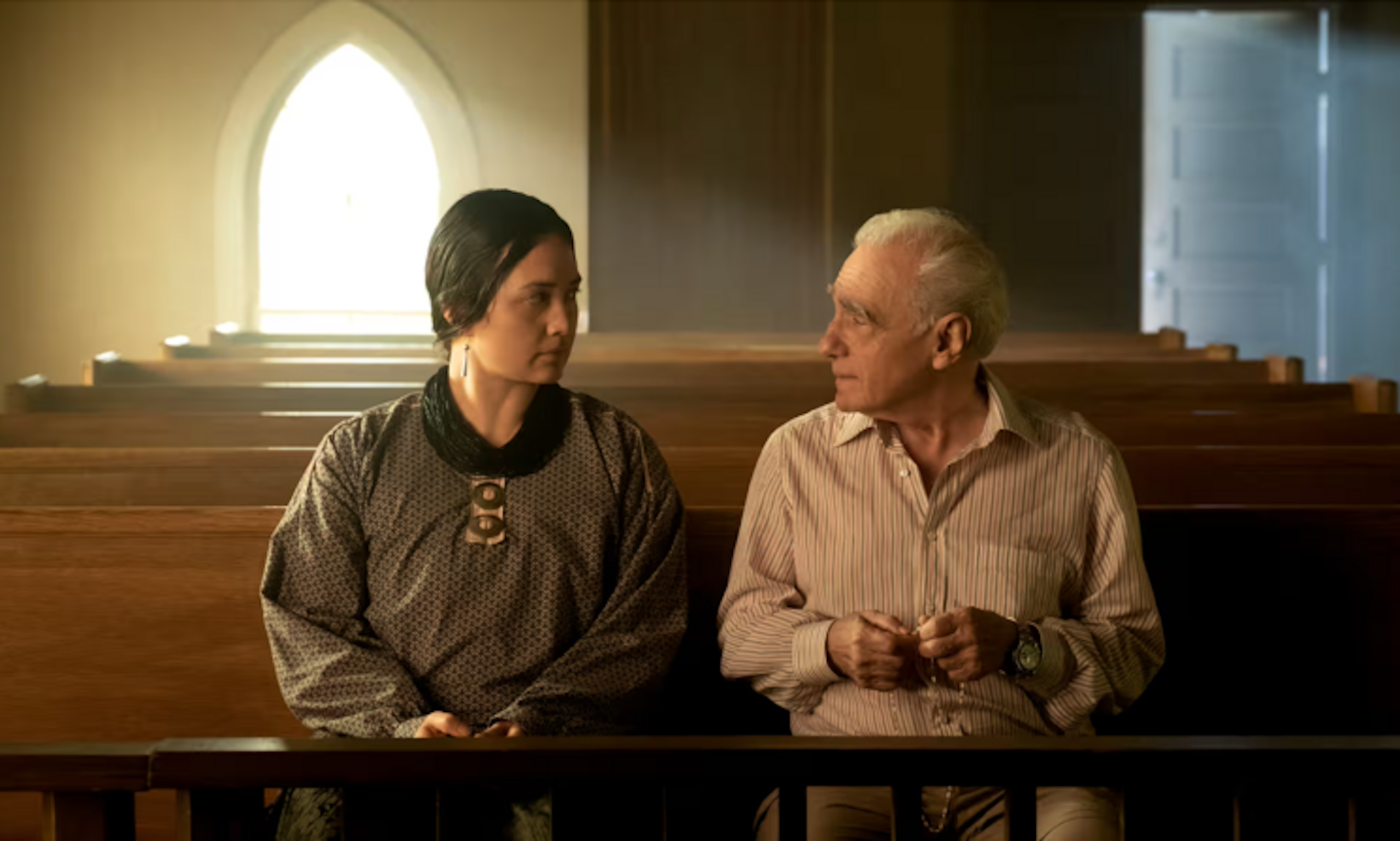 A dark-haired woman in a dark dress and a white-haired man in a button-down shirt sit looking at each other seriously in a church pew.