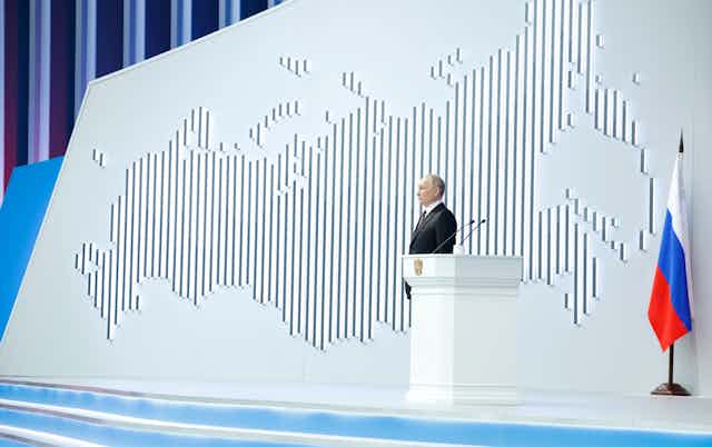 Vladimir Putin stands on a stage in front of a map of the Russian landmass
