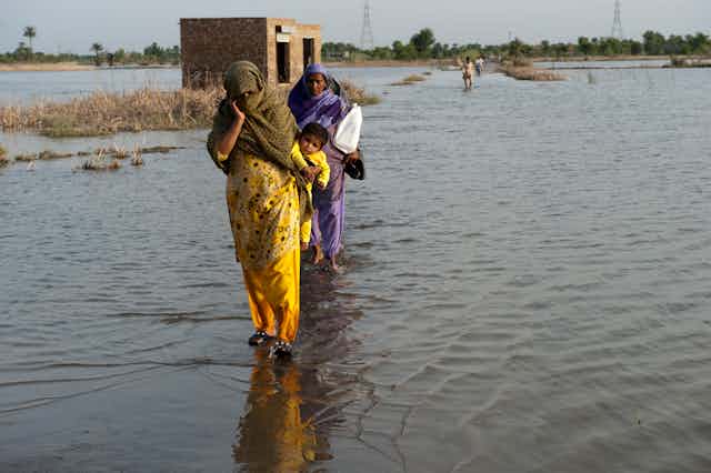 Two women in colourful yellow and purple saris, one with baby on back, walk towards camera on flooded land in Pakistan, trees in distant background. 