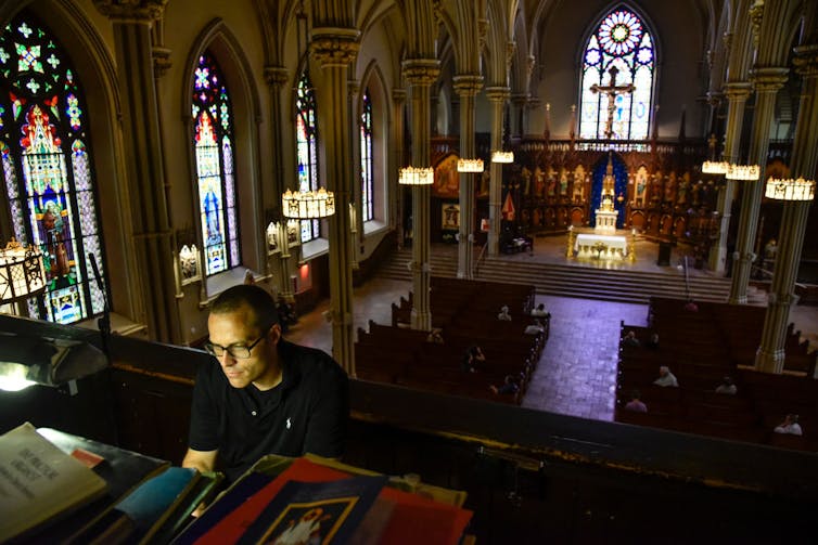 The view of a sanctuary with stained-glass windows, seen from above with a man playing the organ in the foreground.