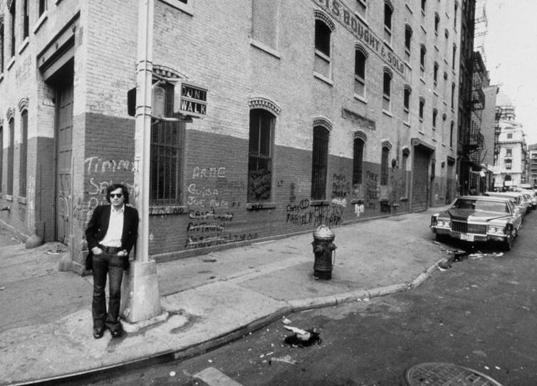 A black and white photo of a man in a jacket and sunglasses leaning against a lamppost on a street with graffiti.