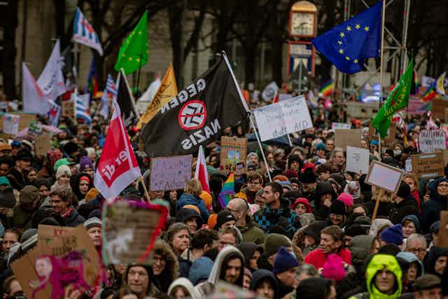 A big crowd of people wear jackets and hold flags, including the blue EU flag and a black one that has the Nazi swastika crossed out.