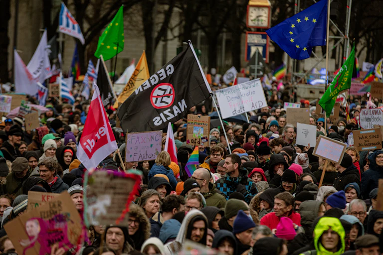 A big crowd of people wear jackets and hold flags, including the blue EU flag and a black one that has the Nazi swastika crossed out.