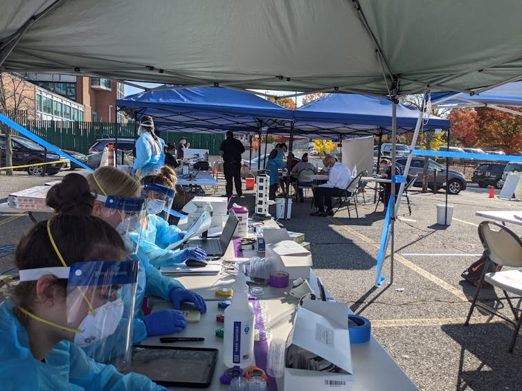 Researchers wearing masks and other protective gear sit under tents collecting samples during a COVID-19 testing study.