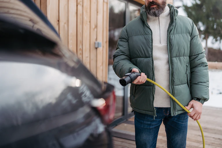 A man wearing a winter coat and glasses stands behind a car holding an electric vehicle charger; in the background there is snow.