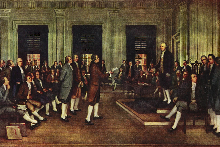 Men in formal 18th century dress stand and sit in a formal meeting room.