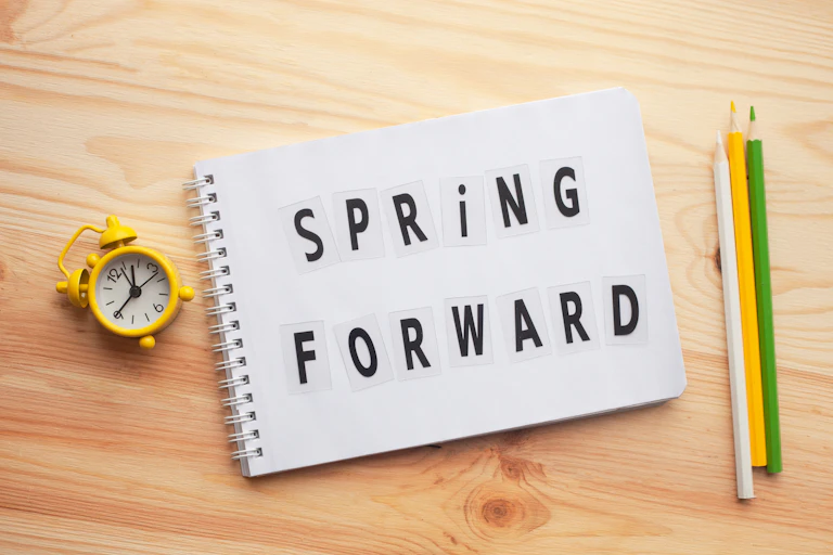 A yellow retro alarm clock, a notepad with the text "Spring Forward" and three pencils lying together on a wooden table.