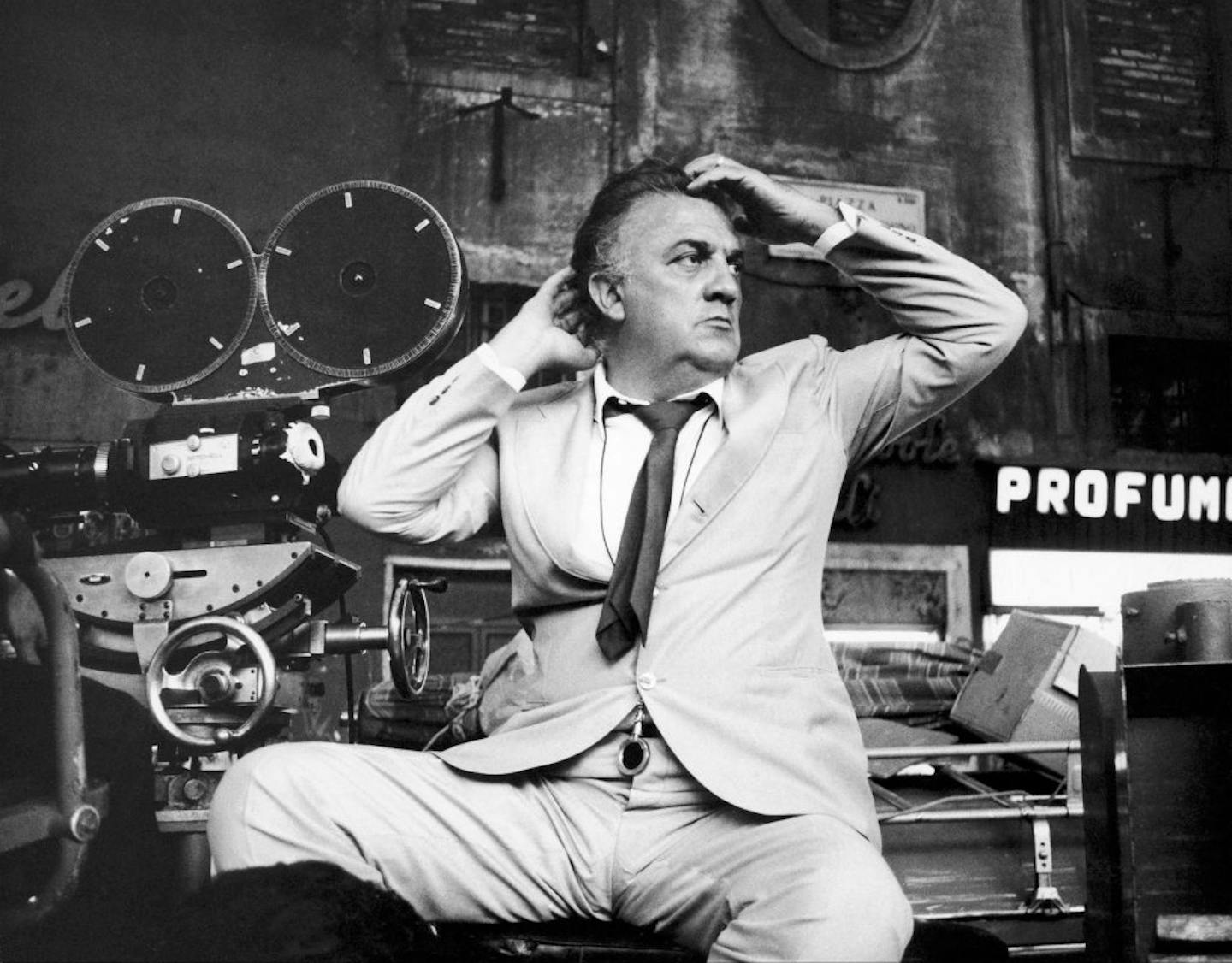 Black and white photo of a middle-aged man running his hands through his hair while sitting in a chair next to a large camera.