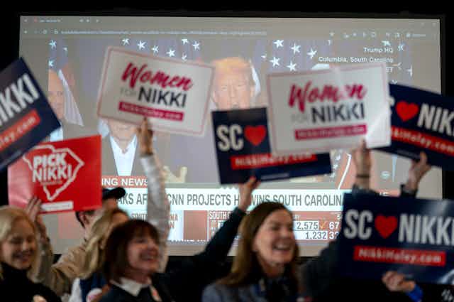Women holding up signs say 'Women for NIKKI' in front of a tv screen with Donald Trump on it. 
