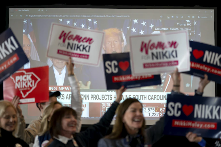 Women holding up signs say 'Women for NIKKI' in front of a tv screen with Donald Trump on it.