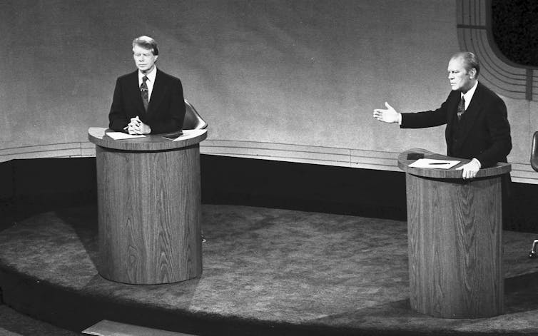 Two men in suits on a stage standing behind individual lecterns.