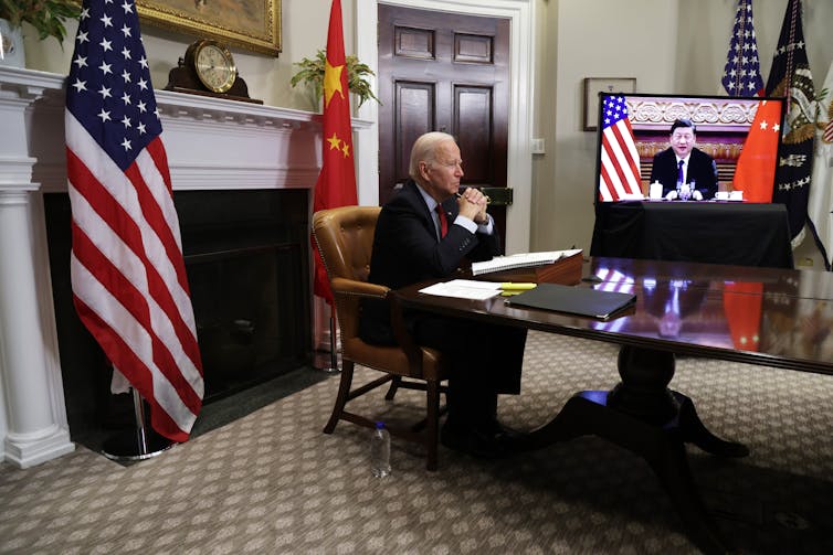 Un hombre sentado se sienta.  escritorio mientras se ve a otro hombre en una pantalla de televisión.