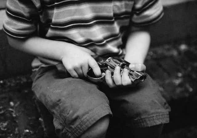 A boy in a striped shirt and shorts holds a revolver. His face is not shown.