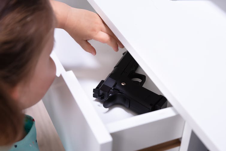 A girl reaches for a black handgun in a white drawer.