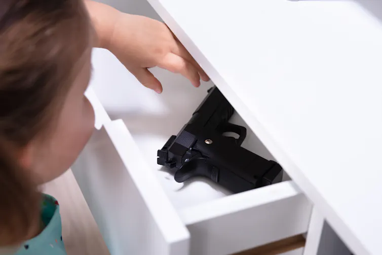 A girl reaches for a black handgun in a white drawer.