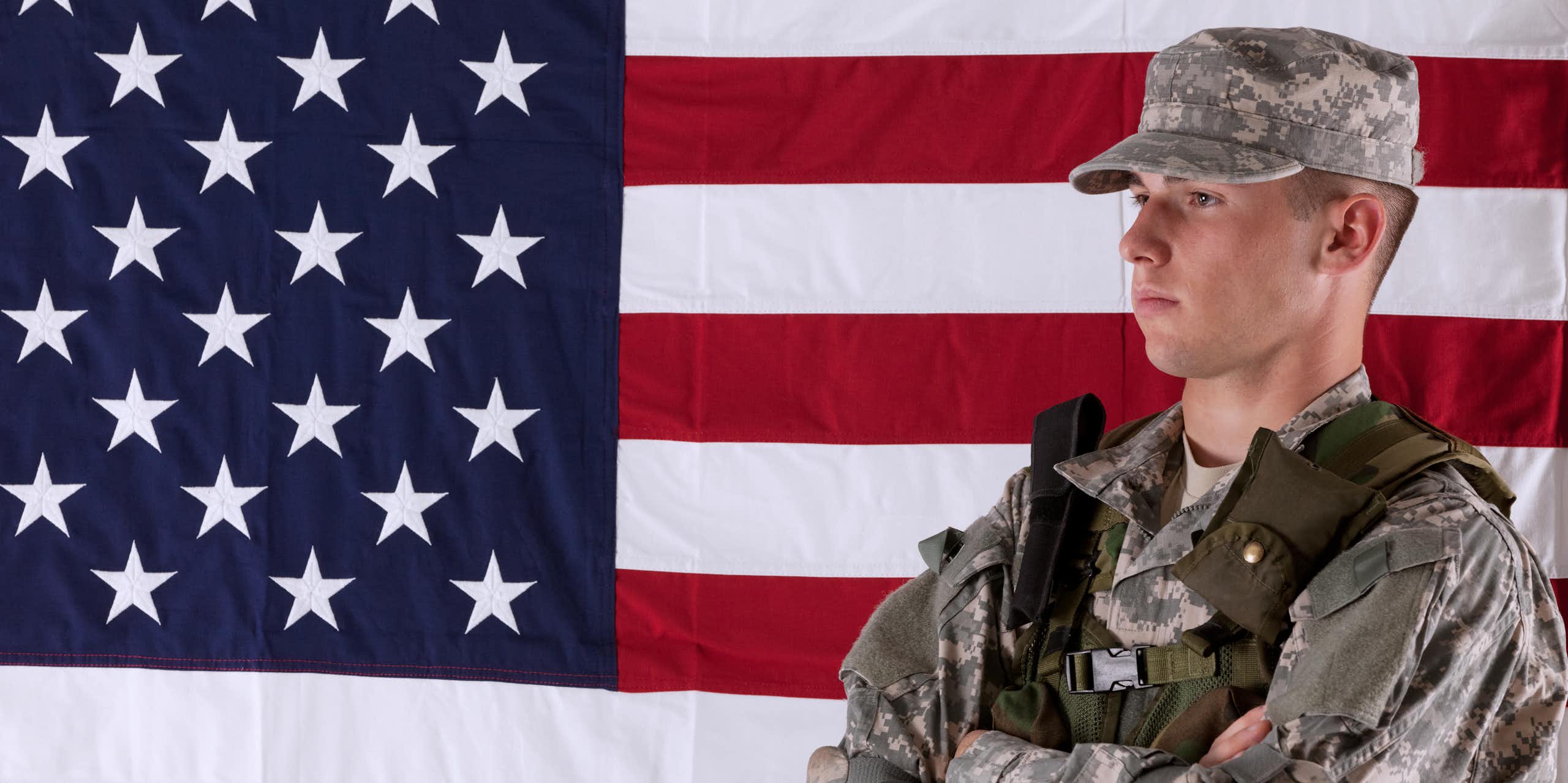 A solider in camouflage stands in front of the right side of an American flag.