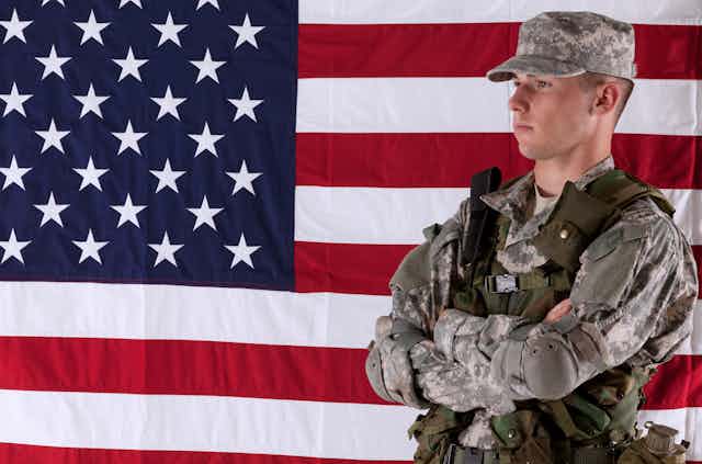 A solider in camouflage stands in front of the right side of an American flag.