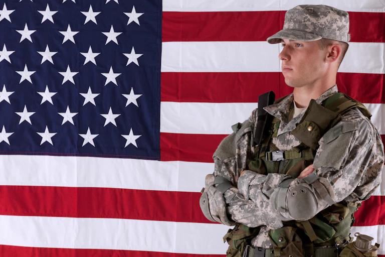 A solider in camouflage stands in front of the right side of an American flag.