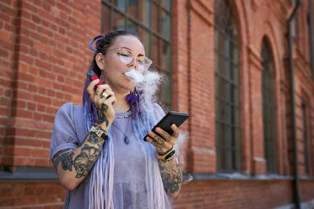 Young woman vaping, holding smartphone, outside redbrick building