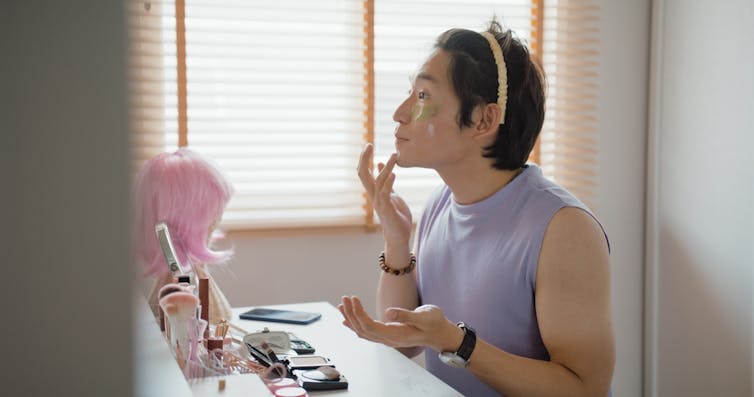 A young Asian man applies makeup at a cluttered vanity.