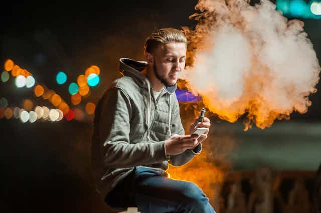 A young man vaping outdoors at night, while looking at his phone.