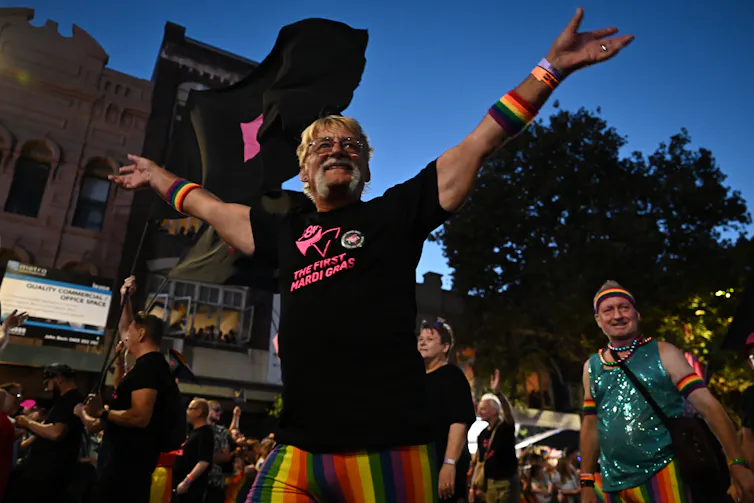 A man in a black t-shirt and rainbow pants walks with this hands in the air