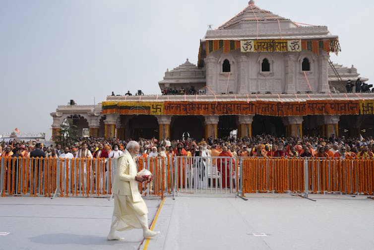 Un hombre camina con una túnica blanca delante de personas vestidas de naranja y un templo.