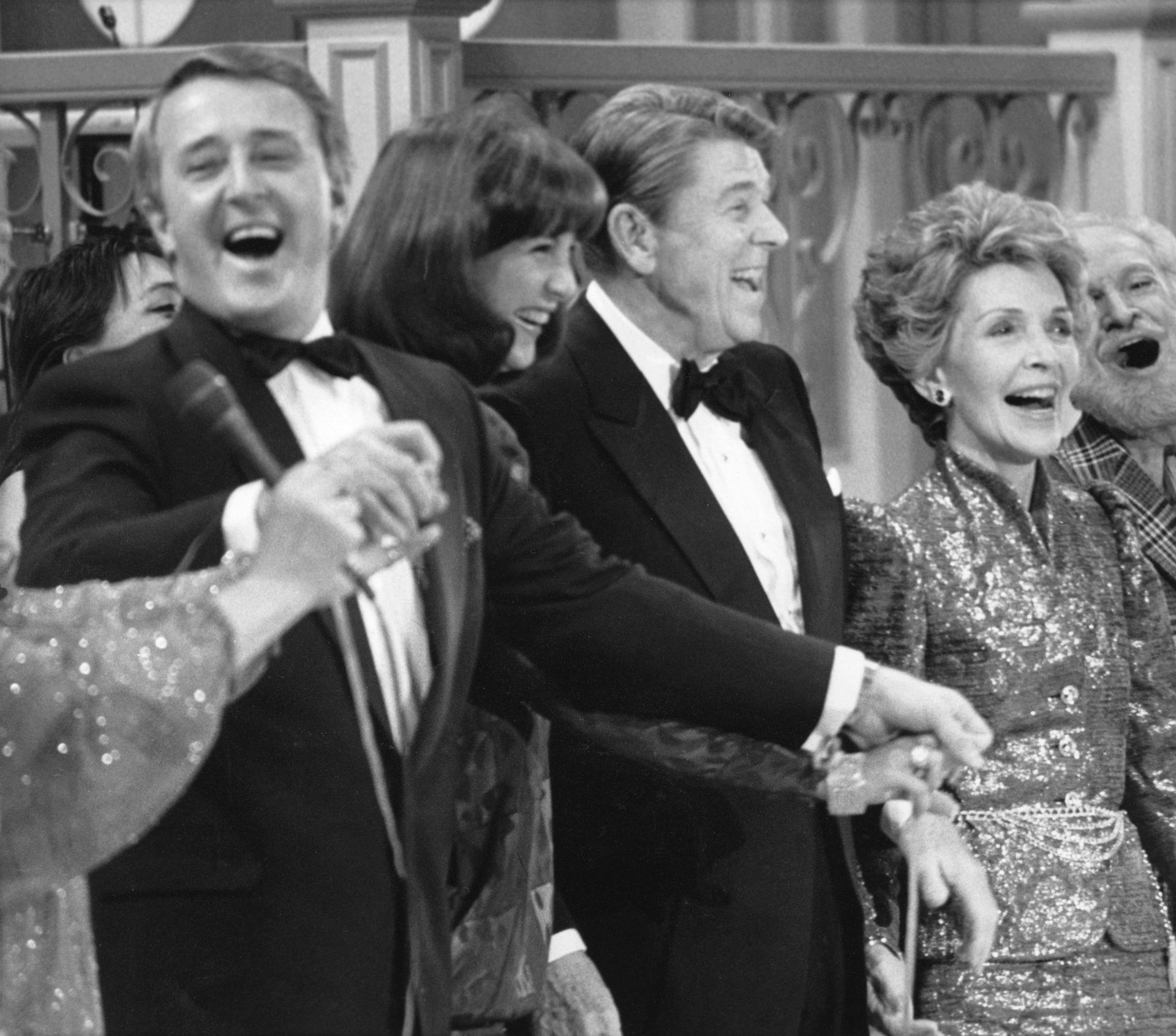 politicians are in tuxedos and their wives in gowns as they grasp hands and sing on a stage