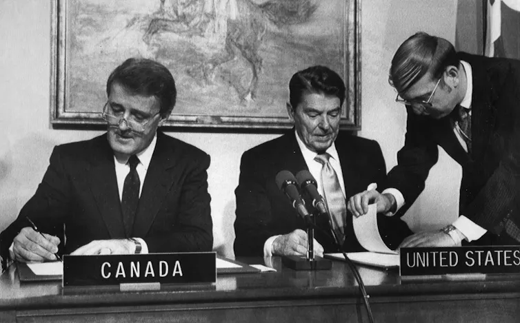 Mulroney and Reagan sit at a desk and sign documents