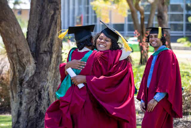 Two people in graduation gowns embrace. A third person in graduation gown looks on, smiling. 