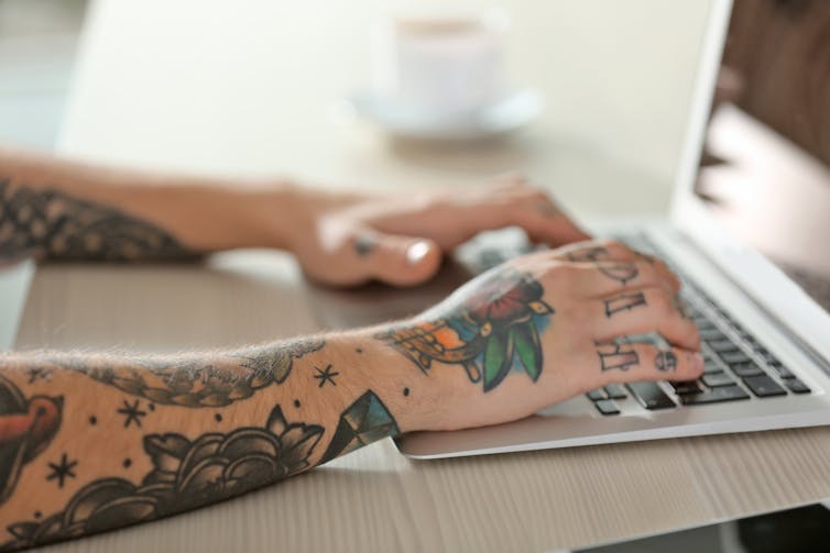Young person with tattoos on arms sitting at desk using laptop