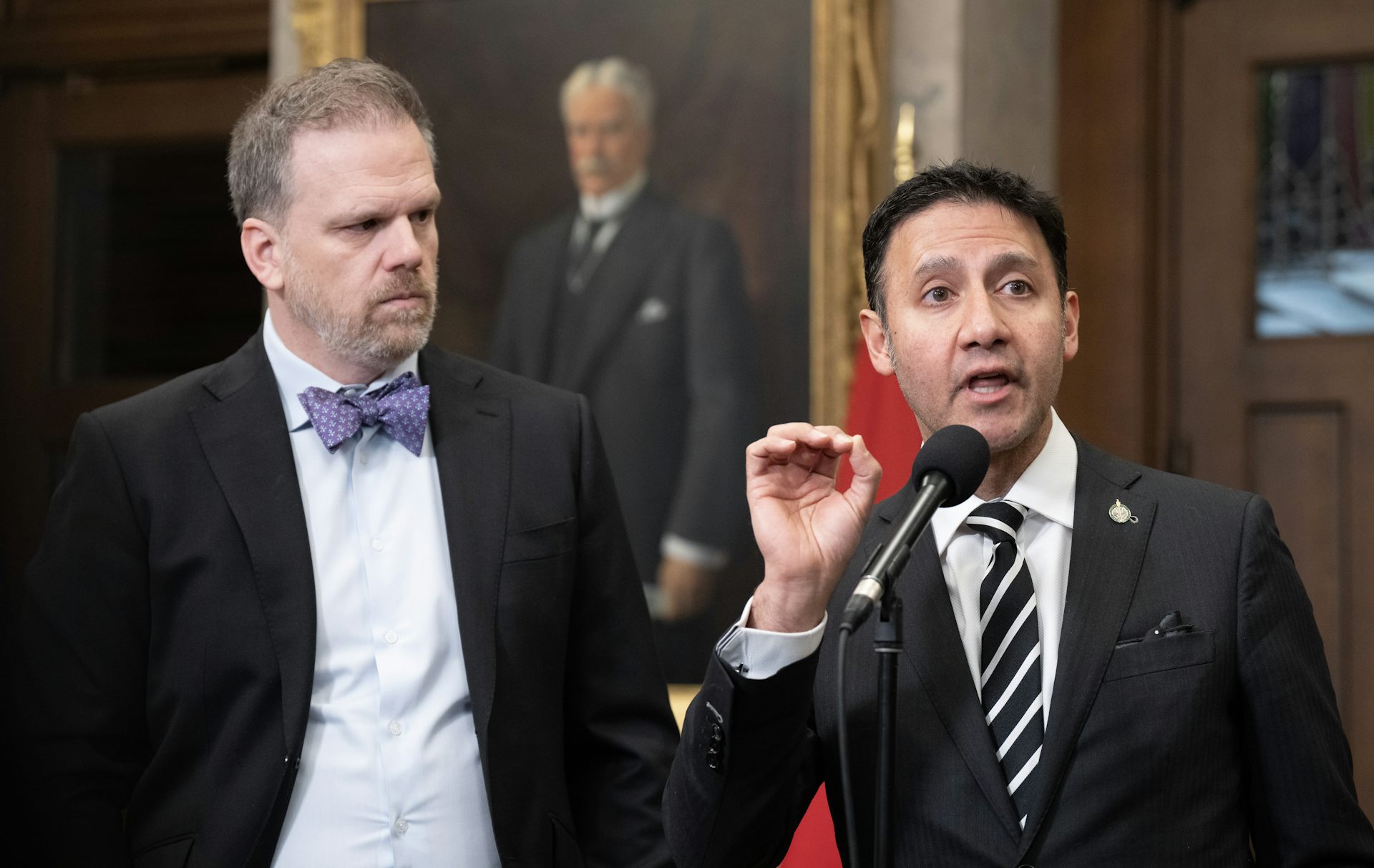 A man in a dark suit at a microphone and another man wearing a bow tie