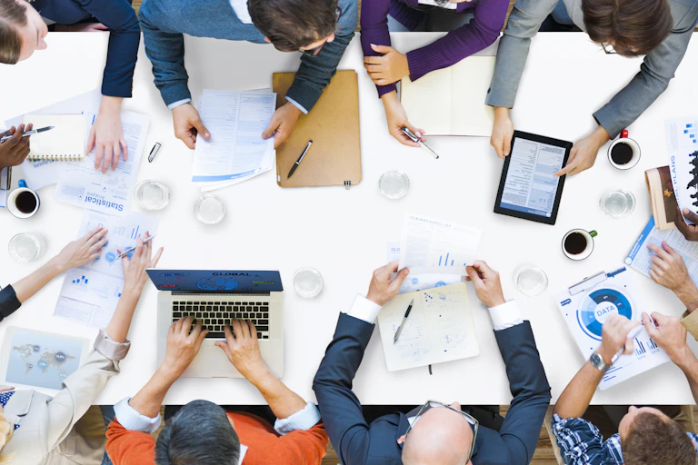 Aerial shot looking down over long boardroom table with people on laptops, drinking coffee, shuffling papers, mix of women, men, ethnic minorities