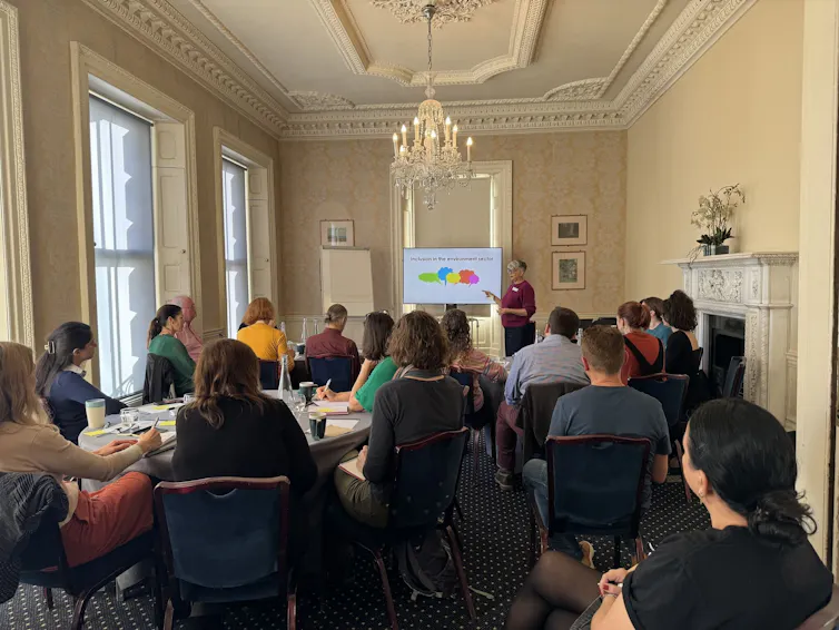 20 or so people sat in white-walled room on chairs, backs facing camera, listening to woman by presentation screen at far end of room, chandelier on ceiling