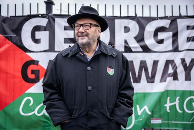 George Galloway, wearing his signature fedora, stands in front of a campaign sign mimicking the Palestinian flag, that reads George Galloway Campaign HQ