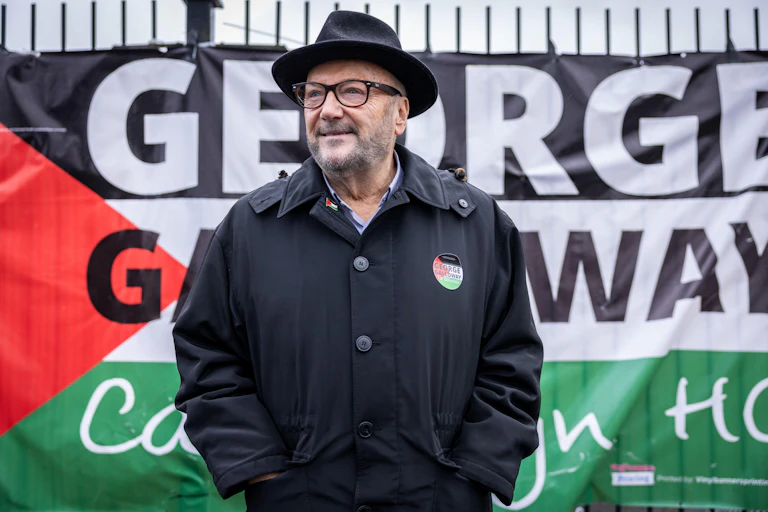 George Galloway, wearing his signature fedora, stands in front of a campaign sign mimicking the Palestinian flag, that reads George Galloway Campaign HQ