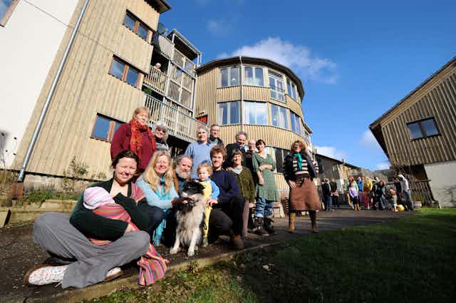 A large group of people and pets stand and sit in front of modern looking buildings. 