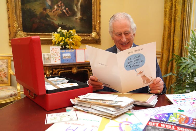 King Charles sits at a desk reading cards from well wishers. He is laughing at a card with a drawing of a dog that says 'at least you don't have to wear a cone!'