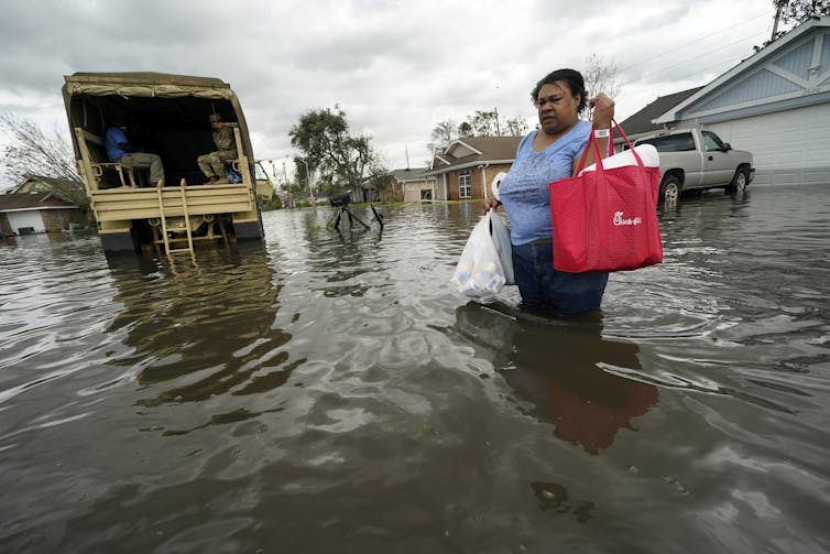 A woman walking in thigh-deep water crosses a road carrying a large bag. A National Guard truck brought her to the home to retrieve medications four days after the hurricane.