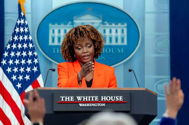 A woman standing at a podium gestures toward people seated in front of her with hands raised