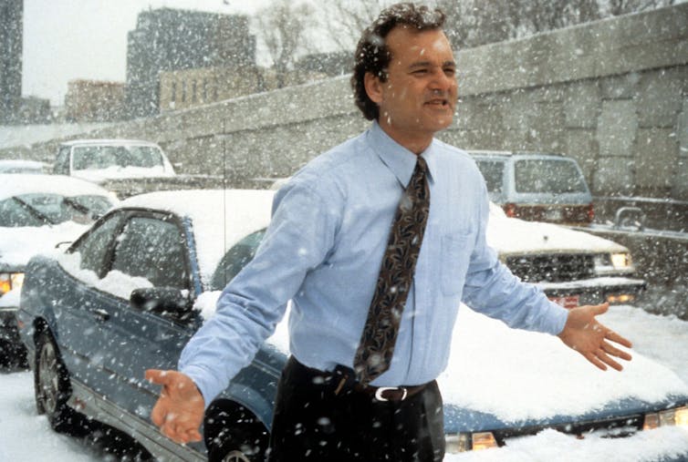 A dark-haired main in a blue shirt and dark tie runs through a snowy street with his arms outstretched.