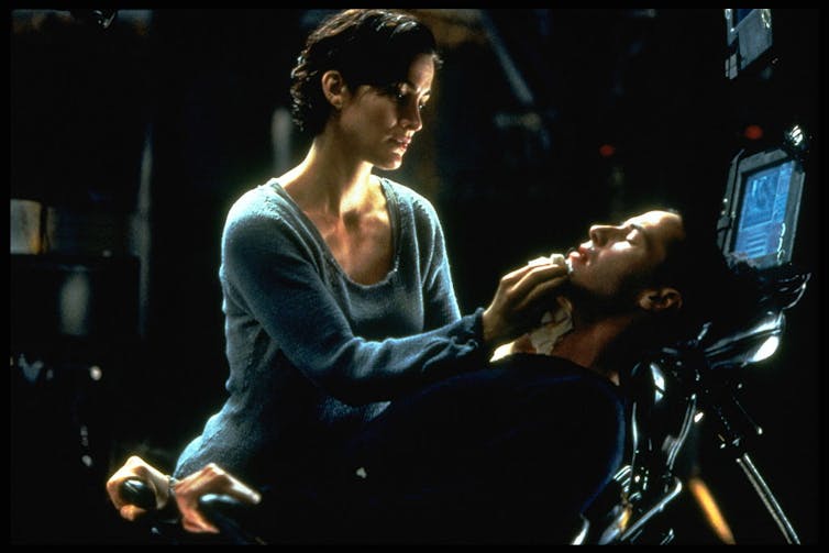 A woman with short hair and a blue shirt touches the chin of a reclining man whose eyes are closed and whose head is almost touching a computer screen.