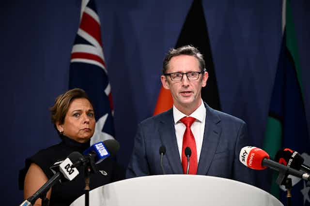 A woman looking stern beside a man in glasses with red tie, both speaking to media microphones in front of Australian and Aboriginal flags