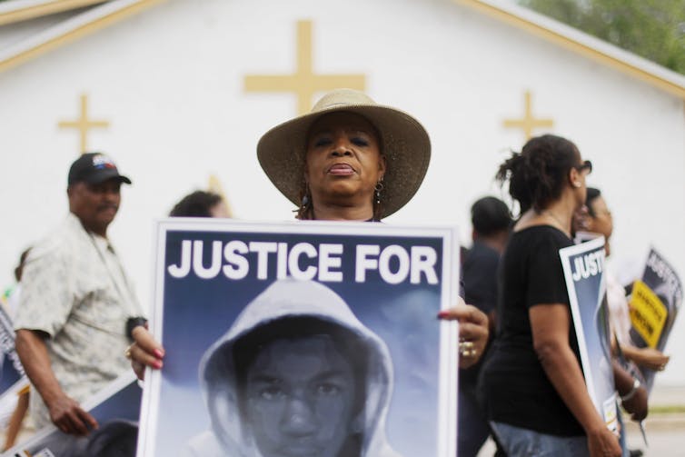 Una mujer negra se encuentra frente a una iglesia y sostiene un cartel de un hombre negro con una sudadera con capucha.
