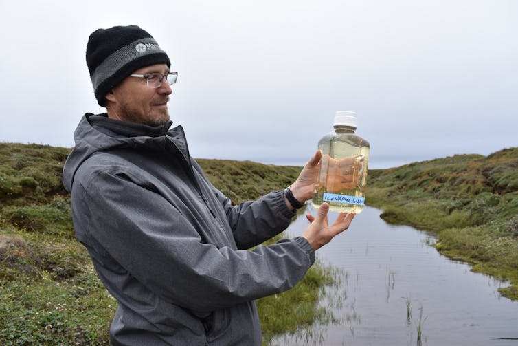 A scientist in a rain jacket and cap holds up a water sample in a jar.